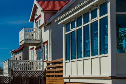 A row of modern homes with large windows and small balconies. The buildings showcase a clean architectural design with white and red detailing. The sky is clear and blue, illuminating the facade of the houses.
