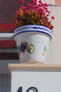 A decorative ceramic pot adorned with colorful floral patterns houses a vibrant plant with small, bright pink flowers. The pot sits atop a white structure, and the background includes red and beige building materials.