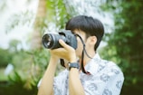 A person with short hair is holding a Canon camera up to their eye, focusing on taking a photograph. They are wearing a patterned shirt and a watch on their left wrist. The background is lush with greenery, suggesting a natural or outdoor setting.