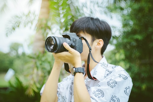 A person with short hair is holding a Canon camera up to their eye, focusing on taking a photograph. They are wearing a patterned shirt and a watch on their left wrist. The background is lush with greenery, suggesting a natural or outdoor setting.
