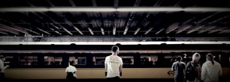 Individuals stand on a train platform, with a train car in the background under an overhead structure. The focal point is a man wearing a white shirt with text, alongside other travelers who appear to be waiting.