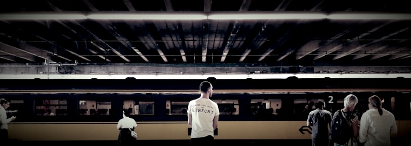 Individuals stand on a train platform, with a train car in the background under an overhead structure. The focal point is a man wearing a white shirt with text, alongside other travelers who appear to be waiting.