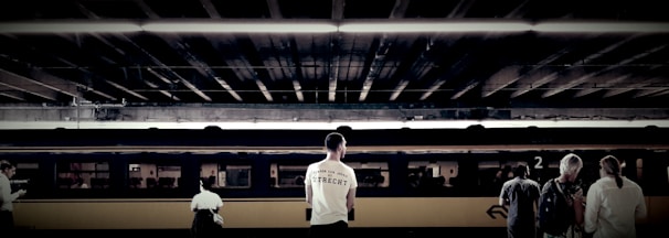 Individuals stand on a train platform, with a train car in the background under an overhead structure. The focal point is a man wearing a white shirt with text, alongside other travelers who appear to be waiting.