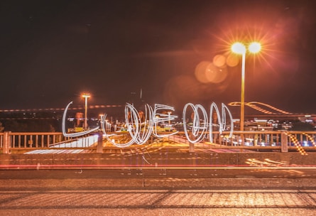 A nighttime scene on a bridge features light trails spelling out 'LIVE ON' from moving lights. Bright streetlights illuminate the surroundings, creating a warm glow and lens flares.