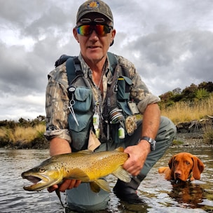 A man dressed in outdoor fishing gear crouches by a river, holding a large brown trout. He wears sunglasses, a cap, and a camo shirt, with various fishing tools attached to his vest. A dog is partially submerged in the water beside him, looking up.