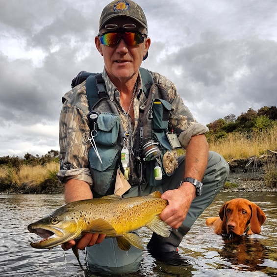 A man dressed in outdoor fishing gear crouches by a river, holding a large brown trout. He wears sunglasses, a cap, and a camo shirt, with various fishing tools attached to his vest. A dog is partially submerged in the water beside him, looking up.