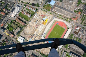 Aerial view of a cityscape featuring a sports stadium with a green field and red running track, adjacent to a large construction site with cranes, surrounded by residential and industrial buildings. A pair of feet in boots is seen standing on a metal beam, adding a sense of height and perspective.