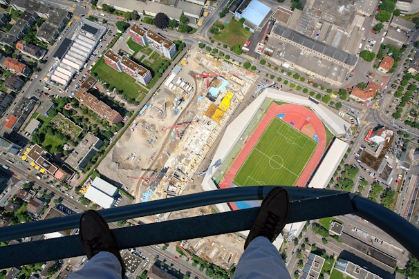 Aerial view of a cityscape featuring a sports stadium with a green field and red running track, adjacent to a large construction site with cranes, surrounded by residential and industrial buildings. A pair of feet in boots is seen standing on a metal beam, adding a sense of height and perspective.