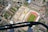 Aerial view of a cityscape featuring a sports stadium with a green field and red running track, adjacent to a large construction site with cranes, surrounded by residential and industrial buildings. A pair of feet in boots is seen standing on a metal beam, adding a sense of height and perspective.