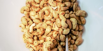 A collection of roasted cashew nuts spread out on a white surface. The nuts vary in size and are golden brown with a glossy texture, indicating they have been seasoned or roasted. A metallic spoon is partially visible among the nuts.