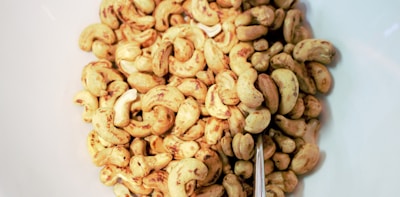 A collection of roasted cashew nuts spread out on a white surface. The nuts vary in size and are golden brown with a glossy texture, indicating they have been seasoned or roasted. A metallic spoon is partially visible among the nuts.