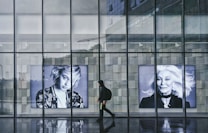 A person dressed in a black hoodie walks past a glass building façade featuring large black-and-white portraits of two women with striking expressions. The ground is reflective, suggesting wet conditions. The glass panels also reveal a reflection of the urban environment in the background.