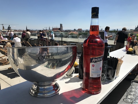 A rooftop setting features a large silver champagne bowl with the brand Cattier visible on it. Next to it is a large bottle of Aperol, an orange-red Italian aperitif. Various bar items like glasses and drink mixers are arranged on the counter. In the background, people are sitting and standing, enjoying the sunny day with a cityscape view.