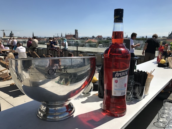 A rooftop setting features a large silver champagne bowl with the brand Cattier visible on it. Next to it is a large bottle of Aperol, an orange-red Italian aperitif. Various bar items like glasses and drink mixers are arranged on the counter. In the background, people are sitting and standing, enjoying the sunny day with a cityscape view.