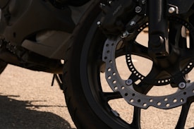 A close-up of a motorcycle wheel featuring detailed view of the brake disc and tire tread. The image shows the metallic texture of the disc with several holes for heat dissipation. The surrounding tire appears robust and well-treaded. Shadows and light play on the surface of the wheel, emphasizing its mechanical components.