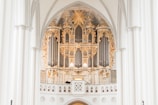 An ornate pipe organ set within a grand architectural setting, featuring intricate gold and white decorative elements. The structure is framed by large arched columns, with elaborate carvings and decorative flourishes surrounding the pipes. The overall design is elevated, emphasizing the craftsmanship with a harmonious blend of classical ornamentation.