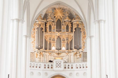 An ornate pipe organ set within a grand architectural setting, featuring intricate gold and white decorative elements. The structure is framed by large arched columns, with elaborate carvings and decorative flourishes surrounding the pipes. The overall design is elevated, emphasizing the craftsmanship with a harmonious blend of classical ornamentation.