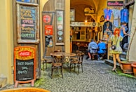 An inviting alleyway café with chalkboard menus displaying drinks like Vinea, Kofola, and coffee. The cobblestone path leads to a cozy interior pub with wooden tables and chairs. Decorative elements include a mannequin dressed in retro fashion and various vintage-style signs. Two people are seated inside, engaged in conversation. Bright, patterned clothing is displayed on racks, and several potted plants add a touch of greenery.