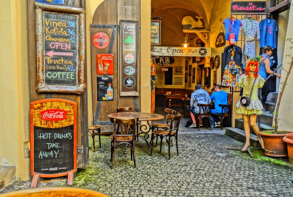 An inviting alleyway café with chalkboard menus displaying drinks like Vinea, Kofola, and coffee. The cobblestone path leads to a cozy interior pub with wooden tables and chairs. Decorative elements include a mannequin dressed in retro fashion and various vintage-style signs. Two people are seated inside, engaged in conversation. Bright, patterned clothing is displayed on racks, and several potted plants add a touch of greenery.