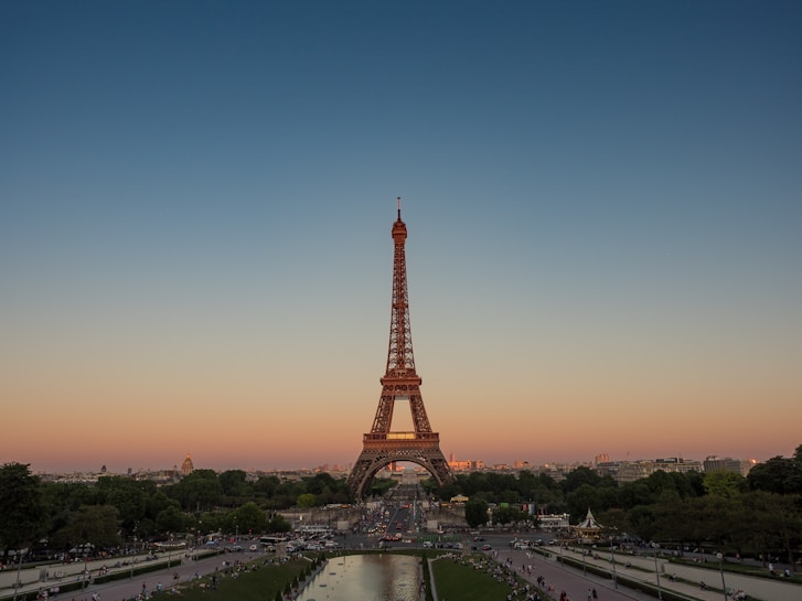 The Eiffel Tower rises majestically against a backdrop of a clear blue to orange gradient sky, marking a stunning sunset. Lush green trees and a park filled with people create a picturesque foreground, while a wide pathway leads up to the iconic structure.