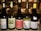 Five wine bottles are arranged on a counter in a dimly lit setting. Each bottle has a distinct label displaying different brands and types of wine, such as Chardonnay, Sauvignon Blanc, and Pinot Gris. In the background, a kitchen environment is visible with chefs in white uniforms working.