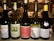 Five wine bottles are arranged on a counter in a dimly lit setting. Each bottle has a distinct label displaying different brands and types of wine, such as Chardonnay, Sauvignon Blanc, and Pinot Gris. In the background, a kitchen environment is visible with chefs in white uniforms working.