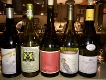 Five wine bottles are arranged on a counter in a dimly lit setting. Each bottle has a distinct label displaying different brands and types of wine, such as Chardonnay, Sauvignon Blanc, and Pinot Gris. In the background, a kitchen environment is visible with chefs in white uniforms working.