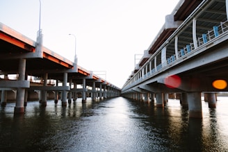 Two parallel concrete bridges span over a body of water, supported by numerous columns. The bridges appear to be partially completed, with construction materials visible on one side. The water below reflects the structure and the sky, creating a sense of depth and perspective.