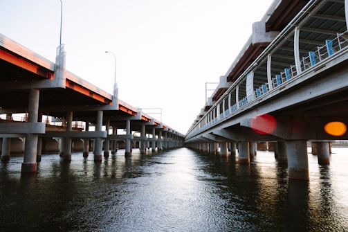 Two parallel concrete bridges span over a body of water, supported by numerous columns. The bridges appear to be partially completed, with construction materials visible on one side. The water below reflects the structure and the sky, creating a sense of depth and perspective.