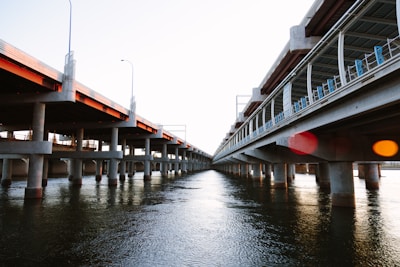 Two parallel concrete bridges span over a body of water, supported by numerous columns. The bridges appear to be partially completed, with construction materials visible on one side. The water below reflects the structure and the sky, creating a sense of depth and perspective.