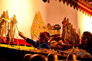 A traditional puppetry performance is taking place, featuring intricately cut and colored shadow puppets. Two performers are visible; one operates the puppets while the other appears to be assisting. The backdrop is adorned with beautifully detailed patterns, and the scene is illuminated with a warm, golden light.