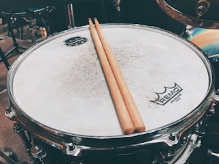 A close-up of a drum with a pair of wooden drumsticks resting on its surface. The drumhead is slightly worn, and logos are visible at the center and edge. The setting includes other drum kit components in the blurred background.