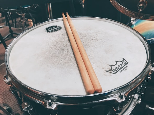 A close-up of a drum with a pair of wooden drumsticks resting on its surface. The drumhead is slightly worn, and logos are visible at the center and edge. The setting includes other drum kit components in the blurred background.