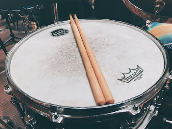 A close-up of a drum with a pair of wooden drumsticks resting on its surface. The drumhead is slightly worn, and logos are visible at the center and edge. The setting includes other drum kit components in the blurred background.