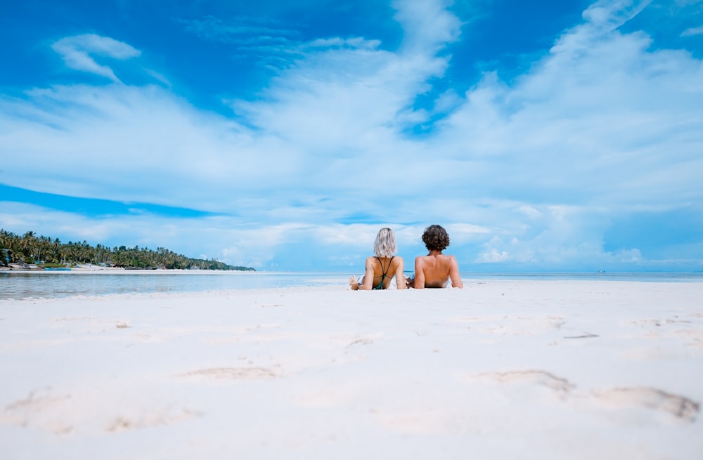 two women lying on white sand facing beach under blue sky