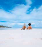 two women lying on white sand facing beach under blue sky