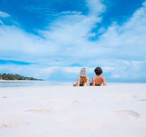 two women lying on white sand facing beach under blue sky