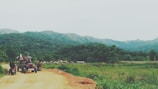A rural landscape featuring a dirt road winding through lush green hills and forests. A tractor with a group of people seated and two motorcycles are visible on the road, moving away from the camera. The distant hills are partially covered in mist, suggesting a serene and tranquil environment.