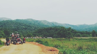 A rural landscape featuring a dirt road winding through lush green hills and forests. A tractor with a group of people seated and two motorcycles are visible on the road, moving away from the camera. The distant hills are partially covered in mist, suggesting a serene and tranquil environment.