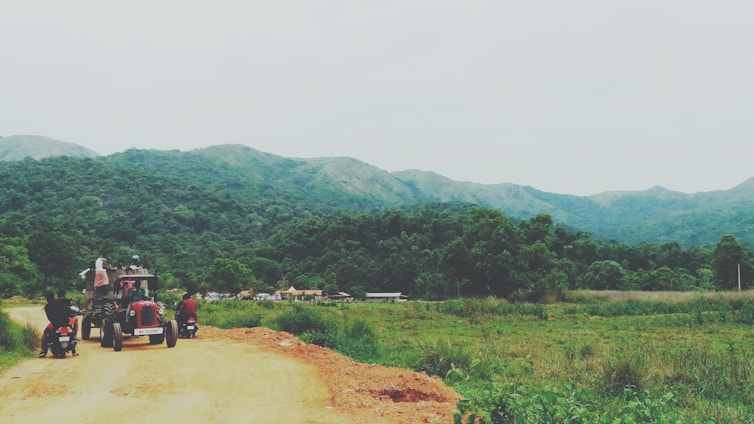 A rural landscape featuring a dirt road winding through lush green hills and forests. A tractor with a group of people seated and two motorcycles are visible on the road, moving away from the camera. The distant hills are partially covered in mist, suggesting a serene and tranquil environment.