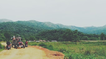 A rural landscape featuring a dirt road winding through lush green hills and forests. A tractor with a group of people seated and two motorcycles are visible on the road, moving away from the camera. The distant hills are partially covered in mist, suggesting a serene and tranquil environment.