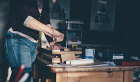 A person is engaged in a woodworking project, using tools to work on several pieces of wood stacked on a table in a workshop. The surroundings include various containers and boxes, with artwork partially visible on the wall. Soft natural light casts shadows, creating a calm and focused atmosphere.