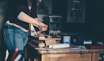 A person is engaged in a woodworking project, using tools to work on several pieces of wood stacked on a table in a workshop. The surroundings include various containers and boxes, with artwork partially visible on the wall. Soft natural light casts shadows, creating a calm and focused atmosphere.