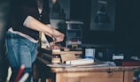 A person is engaged in a woodworking project, using tools to work on several pieces of wood stacked on a table in a workshop. The surroundings include various containers and boxes, with artwork partially visible on the wall. Soft natural light casts shadows, creating a calm and focused atmosphere.