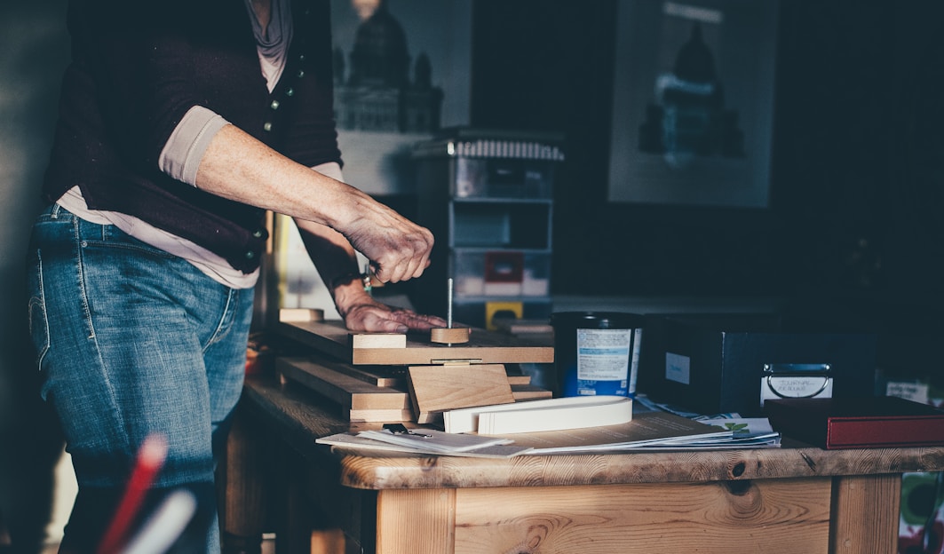 A person is engaged in a woodworking project, using tools to work on several pieces of wood stacked on a table in a workshop. The surroundings include various containers and boxes, with artwork partially visible on the wall. Soft natural light casts shadows, creating a calm and focused atmosphere.