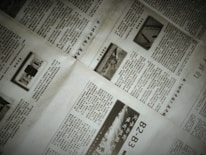 A close-up of a newspaper with multiple pages spread out. The text is written in a non-Latin script, possibly Chinese. There are also images within the articles, including one with the word 'NEWS'. The pages are slightly overlapping, creating an organized yet informal layout.