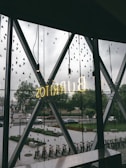 A view through a large window with decorative triangular patterns, revealing an outdoor scene with trees, cars, and bicycles. The word 'BURRITOS' is illuminated in neon, reflecting onto the glass. The setting appears urban, with a street and a bike rack visible.