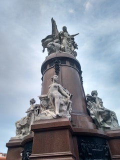 A tall monument featuring elaborate sculptures. The sculptures include several human figures carved in a neoclassical style. The monument's base is dark brown, providing contrast to the light stone figures. The figures appear to be posed symbolically, with one central figure standing upright and others seated around the monument.