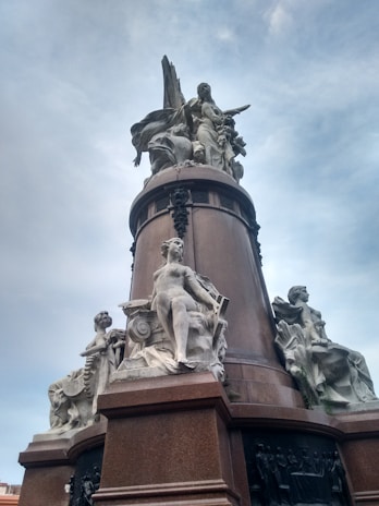 A tall monument featuring elaborate sculptures. The sculptures include several human figures carved in a neoclassical style. The monument's base is dark brown, providing contrast to the light stone figures. The figures appear to be posed symbolically, with one central figure standing upright and others seated around the monument.