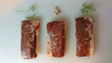 Three raw steaks seasoned with pepper are laid side by side on a light surface. Fresh herbs and pieces of garlic are placed artistically around the steaks for decoration.